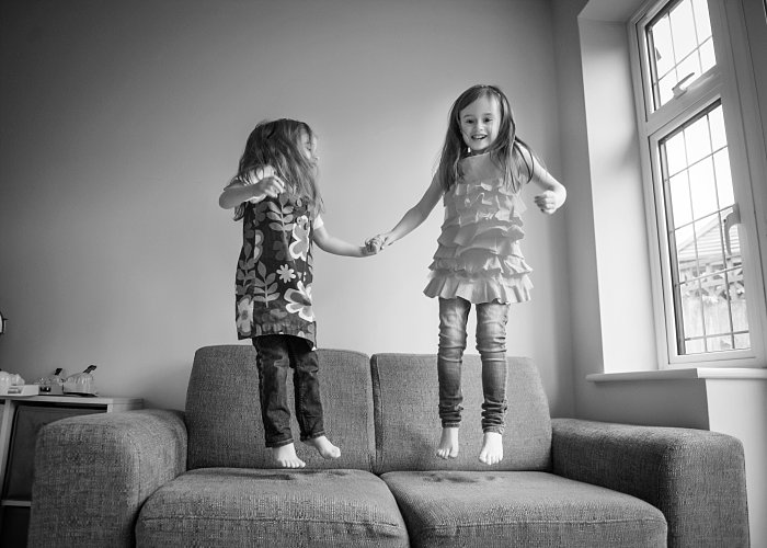 Black and white candid portrait of two young sisters holding hands and jumping on the sofa in their home in Surrey. Captured naturally by Surrey family photographer Maria Cristina Licata.