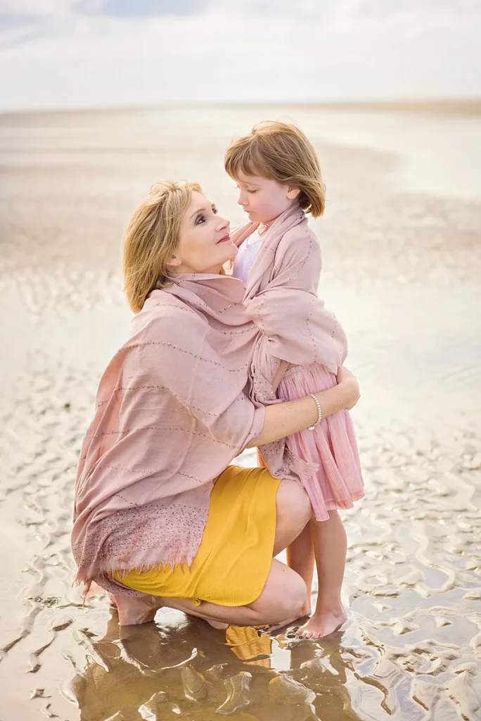 beach portrait of a mum embracing her daughter pink dress under a pink shawl. Captured at West Wittering beach by outdoor family photographer Maria Cristina Licata