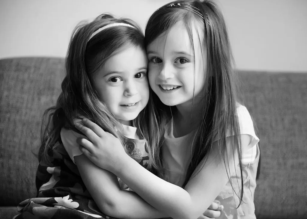 Black and white portrait of two young sisters cuddling and smiling in their home in Surrey. Captured naturally by Surrey family photographer Maria Cristina Licata.