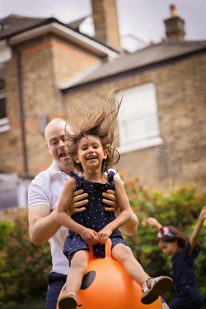 Happy candid family portrait of dad young daughter on a space hopper in their garden in Surrey. Captured by Surrey family photographer Maria Cristina Licata