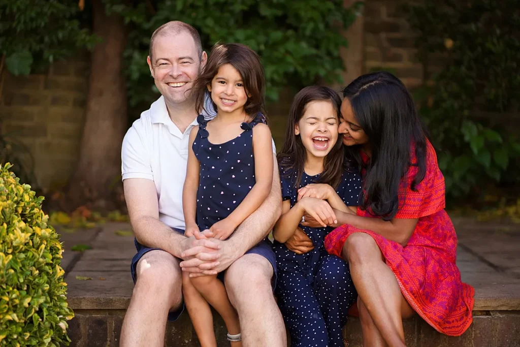 Happy candid family portrait of mum, dad and two young daughters cudddling and laughing in their garden in Surrey. Captured by Surrey family photographer Maria Cristina Licata