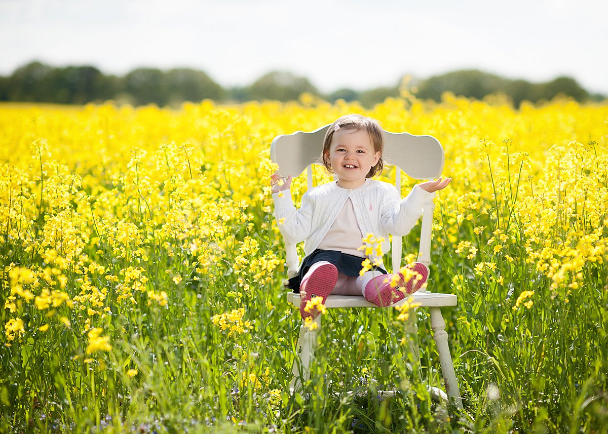 A sunny portrait of a little girl sitting on a chair on the edge of a rape field during a Spring family photoshoot