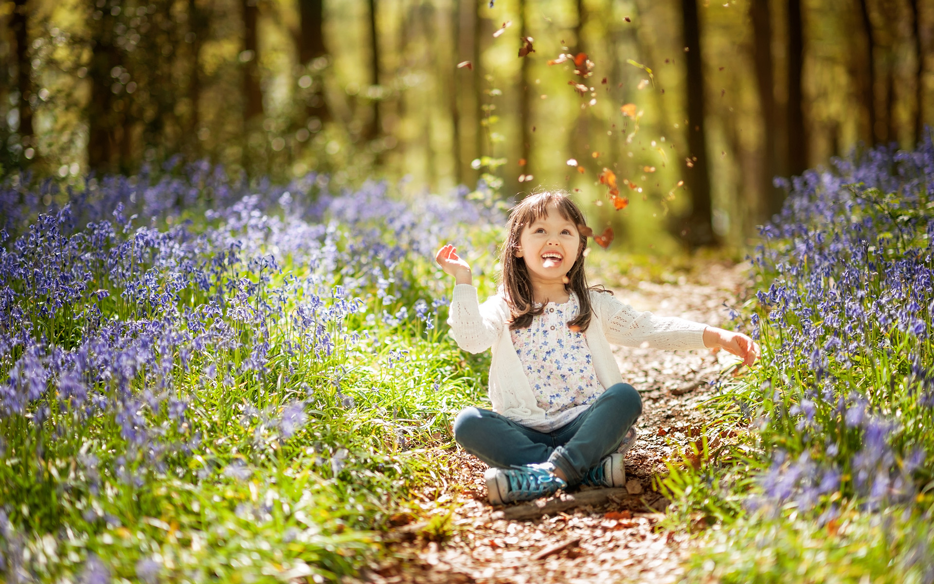 Spring photo of a young girl sitting in a path among the bluebells throwing rusty leaves in the air. Captured by a Hampshire family photographer