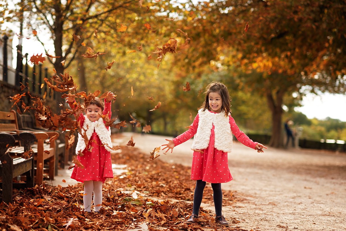 autumn-family-photoshoot_0075 Two young sisters in matching red dresses playing with fallen leaves during an autumn family photoshoot in Richmond – candid seasonal photography by Maria Cristina Licata.