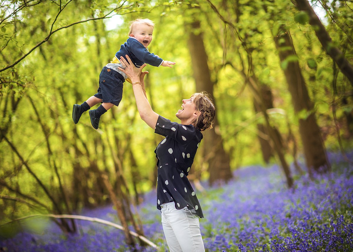 spring-family-photoshoot_0105 Joyful mother lifting her baby high into the air amidst a vibrant bluebell wood during an outdoor family photoshoot in Hampshire – relaxed photography by Maria Cristina Licata.