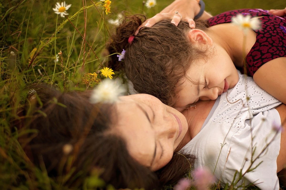 summer-family-photoshoot_0084 Tender moment of mother and daughter lying together among wildflowers during a summer family photoshoot in Surrey – intimate family portrait session by Maria Cristina Licata.