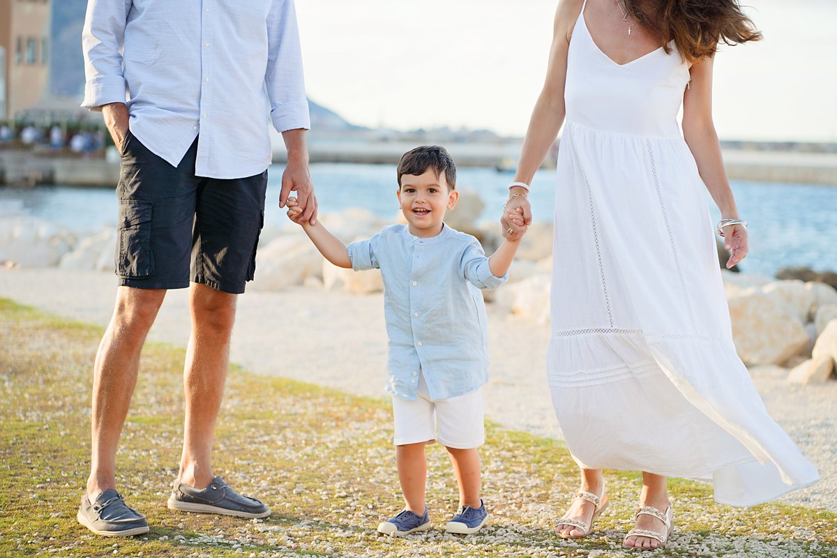 summer-family-photoshoot_0092 Happy family of three walking together by the waterfront, holding hands during a relaxed outdoor portrait session by Maria Cristina Licata.