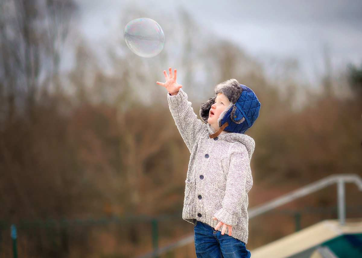 winter-family-portraits_0064 Young child in cozy winter clothing reaching up to catch a soap bubble outdoors – playful moment captured during a family photography session in winter by Maria Cristina Licata