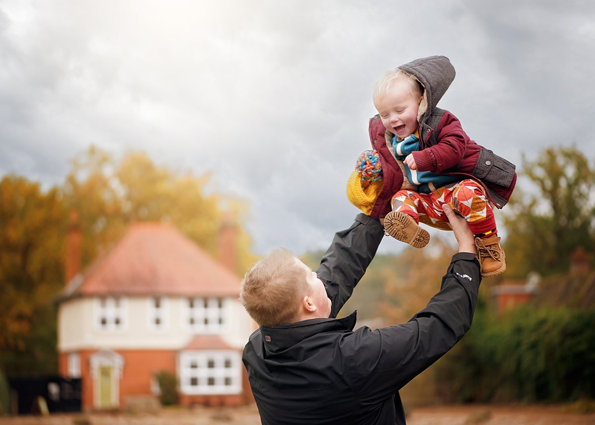 winter-family-portraits_0067 Joyful father lifting his laughing toddler high in the air during a winter family photoshoot at RHS Wisley, capturing pure father-child connection by Maria Cristina Licata.