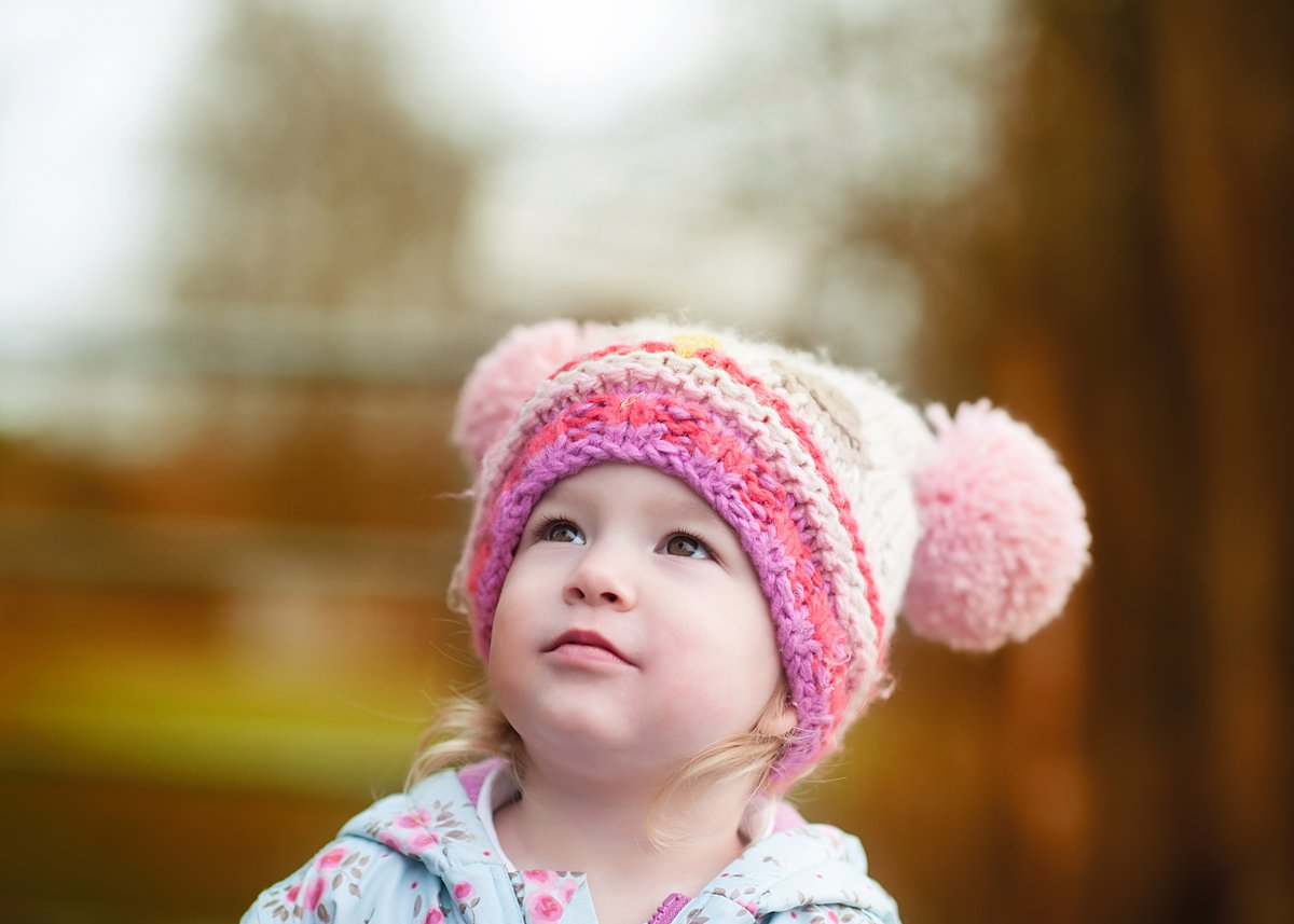 winter-family-portraits_0072 Sweet toddler in a colorful knitted pom-pom hat gazing upward with wonder, capturing innocent childhood curiosity during a family session by Maria Cristina Licata.