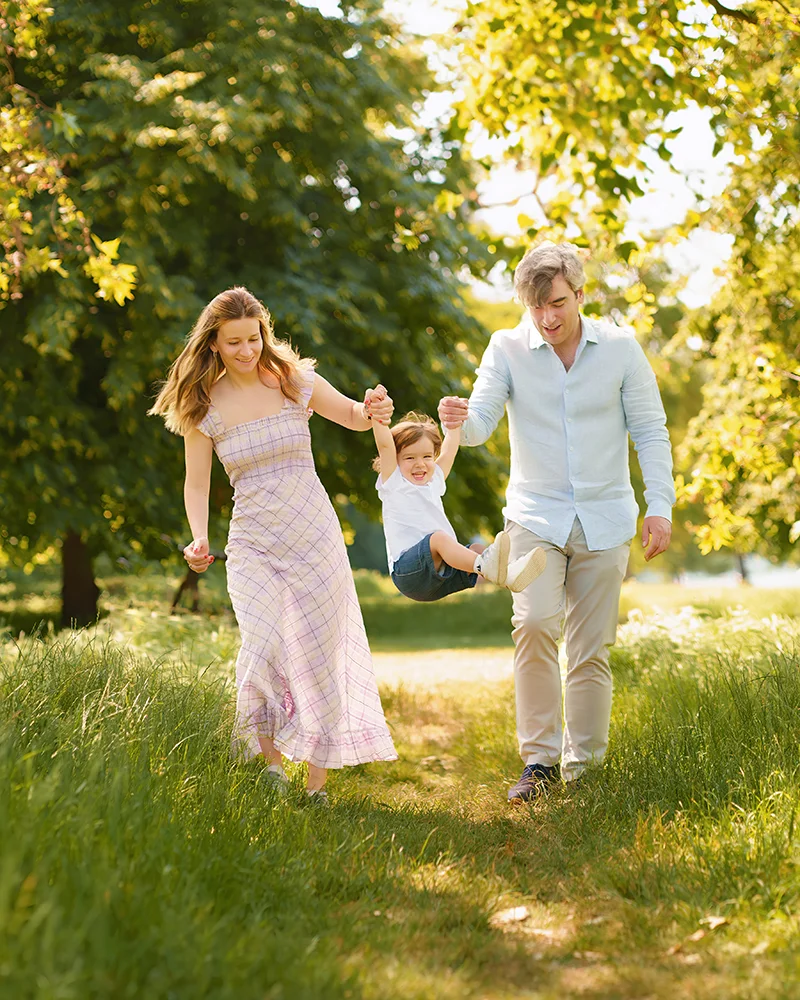 A natural family portrait of mum and dad swinging their little boy in the air in a London park on a sunny summery day. Captured during a photoshoot with family photographer Maria Cristina Licata