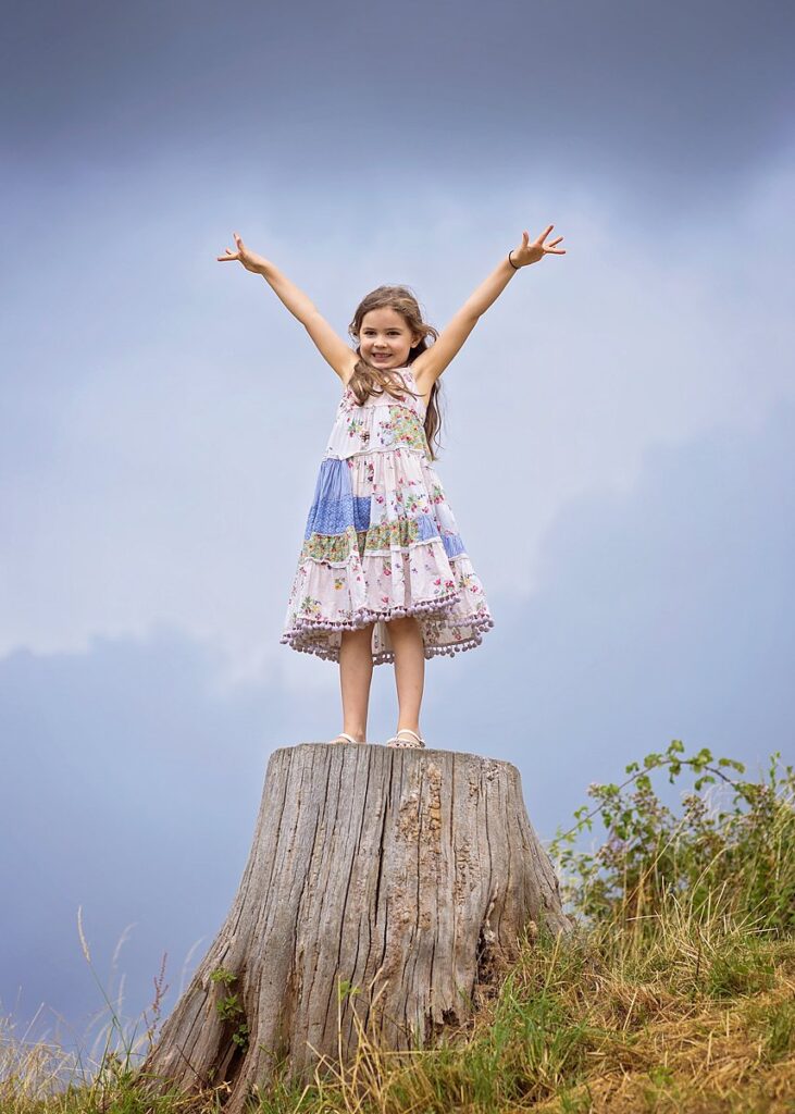 Fun portrait of a girl standing on a tree stump, with a dramatic grey sky in the background. Taken during a family photoshoot at Silchester, near Reading