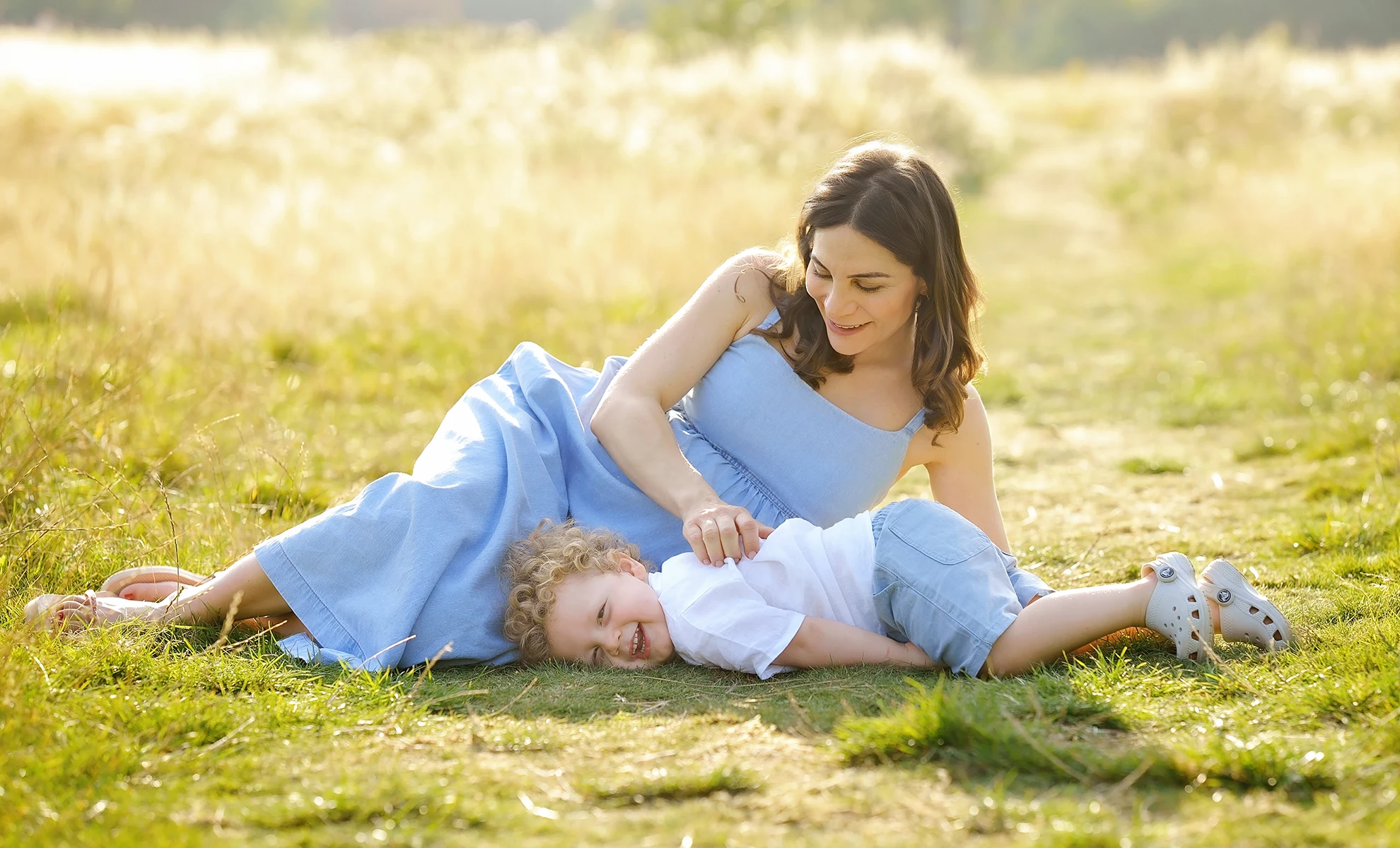 a playful portrait of a mum and toddler son, sitting on the grass at Hampstead Heath in London during a summer family photoshoot at sunset. Captured by leading hampshire family photographer Maria Cristina Licata